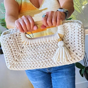 Pode incluir: Bolsa tote creme, feita &agrave; m&atilde;o em croch&ecirc;, com al&ccedil;as de madeira e uma borla creme. A bolsa tem formato retangular e um ponto grosso e texturizado. As al&ccedil;as s&atilde;o acentuadas com um design de croch&ecirc; amarelo e laranja.