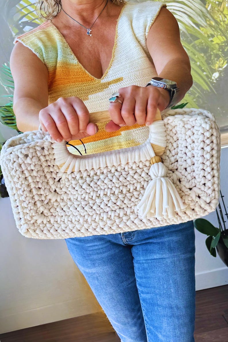 Pode incluir: Bolsa tote creme, feita &agrave; m&atilde;o em croch&ecirc;, com al&ccedil;as de madeira e uma borla creme. A bolsa tem formato retangular e um ponto grosso e texturizado. As al&ccedil;as s&atilde;o acentuadas com um design de croch&ecirc; amarelo e laranja.