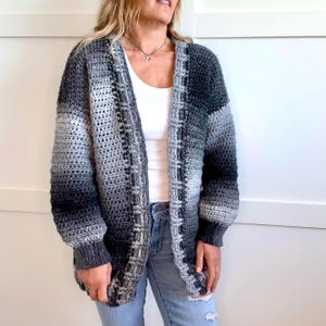 May include: A handmade crochet cardigan in shades of gray, black, and white. The open-front sweater features a textured stitch pattern and long sleeves. The cardigan is a cozy, oversized design.