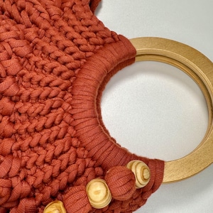 May include: A rust-colored handbag crafted from thick, braided cord featuring a circular light-toned wooden handle. The bag is accented with three round wooden beads and a matching fabric tassel attached to the base of the handle.