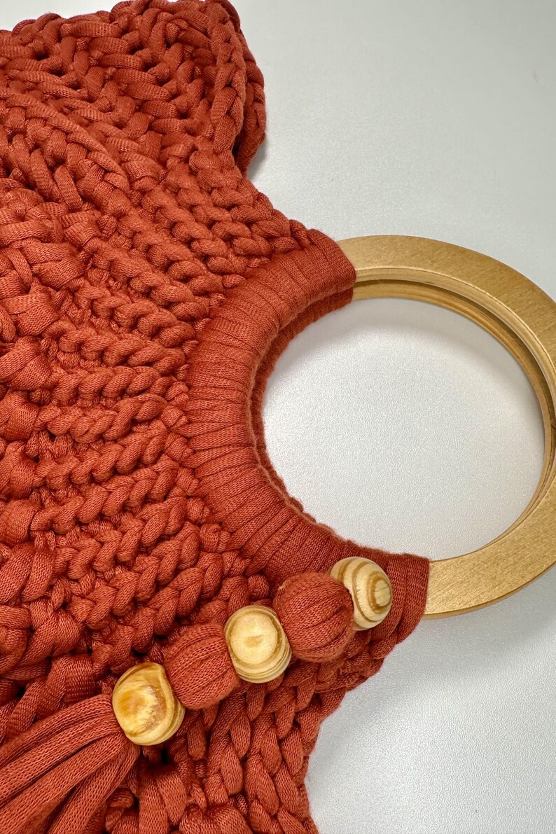 May include: A rust-colored handbag crafted from thick, braided cord featuring a circular light-toned wooden handle. The bag is accented with three round wooden beads and a matching fabric tassel attached to the base of the handle.