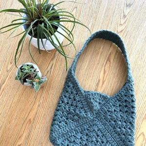 A crochet bag on a table next to 2 small plants. The crochet bag is made with granny squares sewn together, the strap is also crocheted.