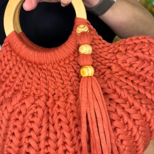 May include: A burnt orange handbag crafted from thick, braided fabric with a round, light-colored wooden handle. The bag features a decorative tassel accent hanging from the handle, adorned with three small, round, light-colored wooden beads.