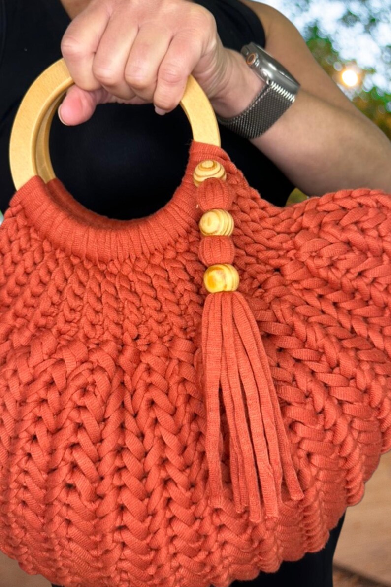 May include: A burnt orange handbag crafted from thick, braided fabric with a round, light-colored wooden handle. The bag features a decorative tassel accent hanging from the handle, adorned with three small, round, light-colored wooden beads.