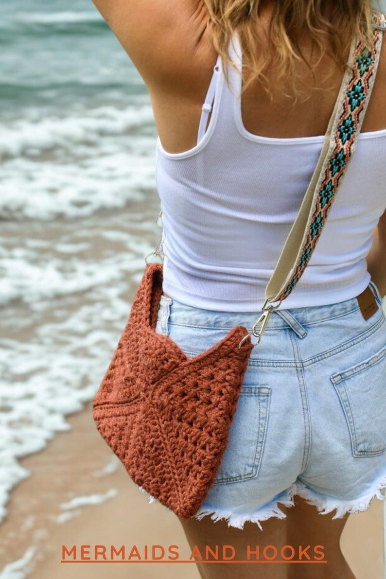 Op de afbeelding: Een roestkleurige gehaakte schoudertas met een gestructureerd geometrisch patroon. De tas heeft een brede, verstelbare riem versierd met een kleurrijk geweven patroon in tinten turquoise, oranje en zwart. De tekst MERMAIDS AND HOOKS is zichtbaar.
