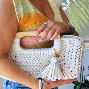 Pode incluir: Bolsa tote de croch&ecirc; creme com al&ccedil;as de madeira e borla. A bolsa apresenta um padr&atilde;o de ponto grosso e uma forma retangular. As al&ccedil;as s&atilde;o de madeira clara. A bolsa &eacute; segurada por uma pessoa que usa um top amarelo e laranja.