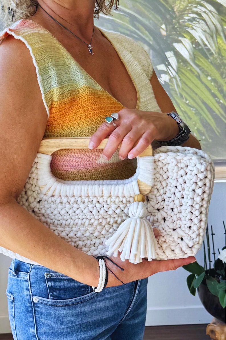Pode incluir: Bolsa tote de croch&ecirc; creme com al&ccedil;as de madeira e borla. A bolsa apresenta um padr&atilde;o de ponto grosso e uma forma retangular. As al&ccedil;as s&atilde;o de madeira clara. A bolsa &eacute; segurada por uma pessoa que usa um top amarelo e laranja.