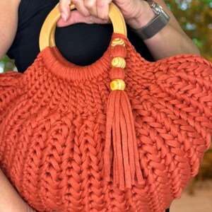 May include: A rust-colored, hand-crocheted handbag featuring a thick, braided yarn texture. The bag includes a round, light-colored wooden handle and a decorative tassel accent attached to the handle with three small, matching wooden beads.