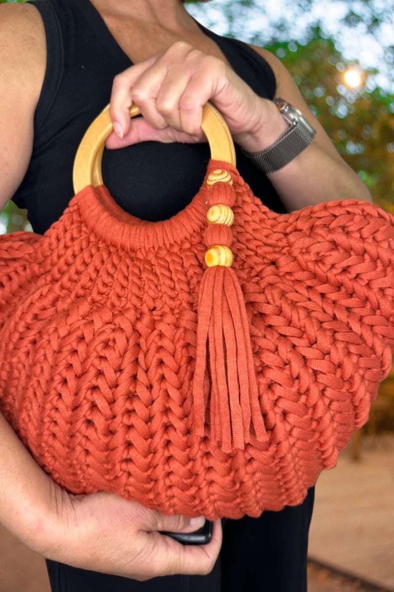 May include: A rust-colored, hand-crocheted handbag featuring a thick, braided yarn texture. The bag includes a round, light-colored wooden handle and a decorative tassel accent attached to the handle with three small, matching wooden beads.