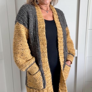 A handmade crochet cardigan in a two-tone design. The upper portion and sleeves are gray, while the body and pockets are mustard yellow. The cardigan features a textured stitch pattern, long sleeves, and two front pockets. The edges are trimmed with a gray border.