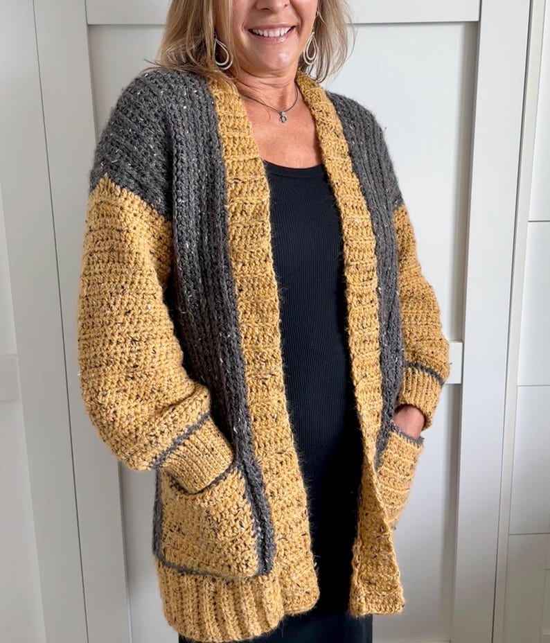 A handmade crochet cardigan in a two-tone design. The upper portion and sleeves are gray, while the body and pockets are mustard yellow. The cardigan features a textured stitch pattern, long sleeves, and two front pockets. The edges are trimmed with a gray border.