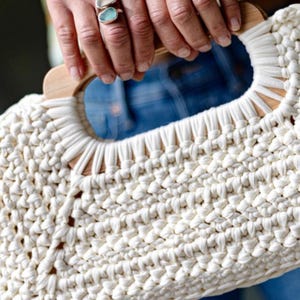Pode incluir: Bolsa de m&atilde;o de macram&eacute; creme com al&ccedil;a de madeira. A bolsa &eacute; feita de algod&atilde;o grosso e tecido, e a al&ccedil;a &eacute; de cor castanho claro. Uma pessoa usa um anel com uma pedra azul.