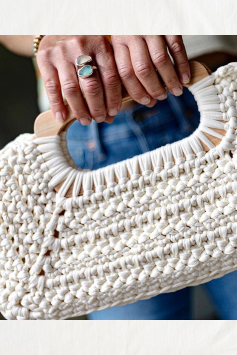 Pode incluir: Bolsa de m&atilde;o de macram&eacute; creme com al&ccedil;a de madeira. A bolsa &eacute; feita de algod&atilde;o grosso e tecido, e a al&ccedil;a &eacute; de cor castanho claro. Uma pessoa usa um anel com uma pedra azul.