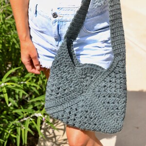 A woman holding a crochet bag that is made with granny squares sewn together, the strap is also crocheted. The woman is wearing denim shorts with a white tank top on a sunny day.