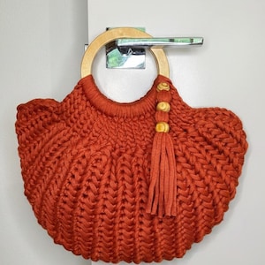 May include: A rust-colored crochet handbag featuring a thick, braided cord texture and a circular light-toned wooden handle. The bag is accented with a matching rust-colored tassel hanging from the handle, which is decorated with three small, round, light-colored wooden beads.