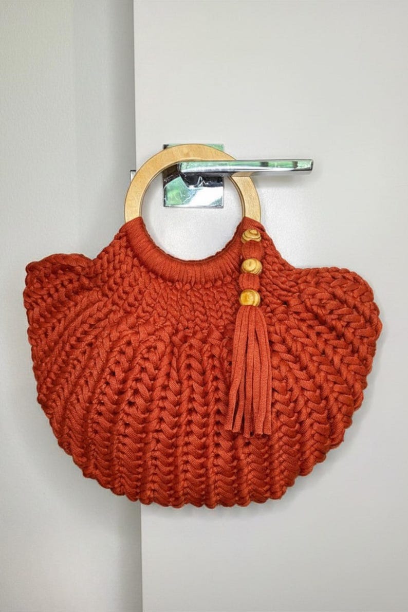May include: A rust-colored crochet handbag featuring a thick, braided cord texture and a circular light-toned wooden handle. The bag is accented with a matching rust-colored tassel hanging from the handle, which is decorated with three small, round, light-colored wooden beads.