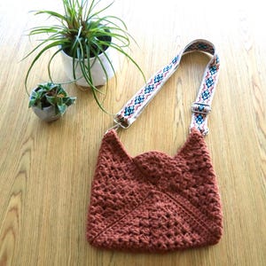 A crossbody crochet bag on a table next to 2 small plants. The crochet bag is made with granny squares sewn together. The colorful strap gives a modern look.