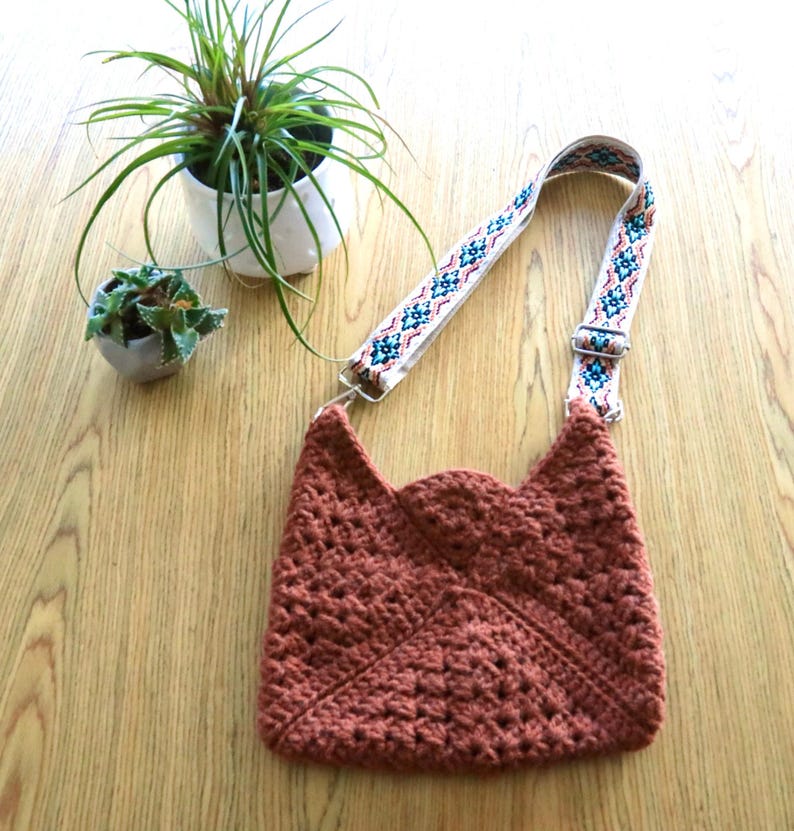 A crossbody crochet bag on a table next to 2 small plants. The crochet bag is made with granny squares sewn together. The colorful strap gives a modern look.