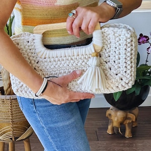 Pode incluir: Bolsa de m&atilde;o de croch&ecirc; creme com al&ccedil;a de madeira e borla. A bolsa tem formato retangular e um padr&atilde;o de ponto grosso e texturizado. A al&ccedil;a &eacute; feita de madeira clara. Uma borla creme pende da lateral da bolsa.