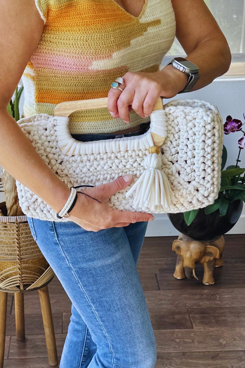 Pode incluir: Bolsa de m&atilde;o de croch&ecirc; creme com al&ccedil;a de madeira e borla. A bolsa tem formato retangular e um padr&atilde;o de ponto grosso e texturizado. A al&ccedil;a &eacute; feita de madeira clara. Uma borla creme pende da lateral da bolsa.