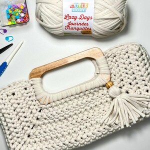 Pode incluir: Bolsa de m&atilde;o de croch&ecirc; creme com al&ccedil;a de madeira e borla. A bolsa est&aacute; ao lado de uma bola de l&atilde;, um recipiente de alfinetes coloridos e ferramentas de croch&ecirc;. O r&oacute;tulo do fio diz "Lazy Days Journ&eacute;es Tranquilles".
