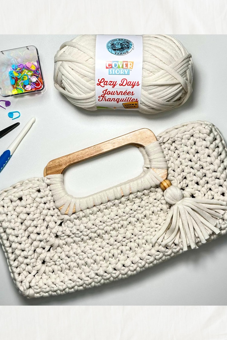 Pode incluir: Bolsa de m&atilde;o de croch&ecirc; creme com al&ccedil;a de madeira e borla. A bolsa est&aacute; ao lado de uma bola de l&atilde;, um recipiente de alfinetes coloridos e ferramentas de croch&ecirc;. O r&oacute;tulo do fio diz "Lazy Days Journ&eacute;es Tranquilles".