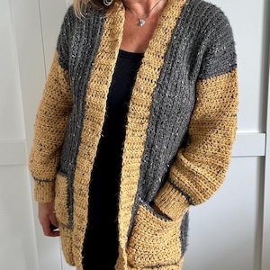 A long, open-front crochet cardigan in a color-block design. The main body is a speckled gray, while the borders, sleeves, and pockets are a solid mustard yellow. The cardigan features a shawl collar and two front pockets.