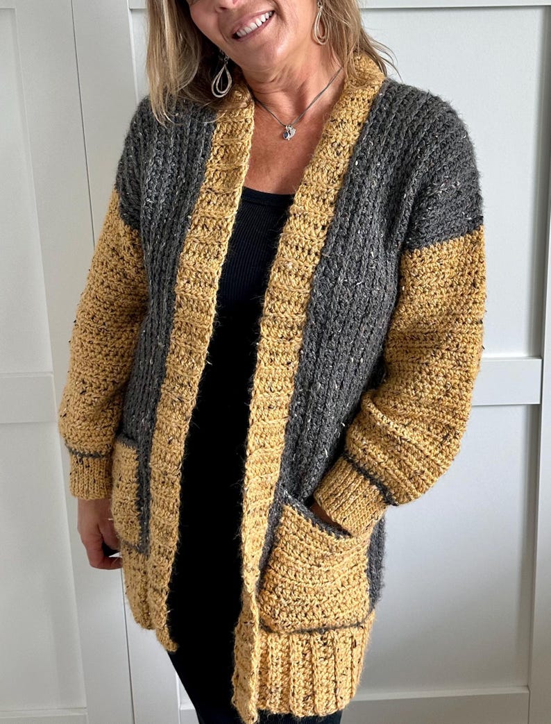 A long, open-front crochet cardigan in a color-block design. The main body is a speckled gray, while the borders, sleeves, and pockets are a solid mustard yellow. The cardigan features a shawl collar and two front pockets.