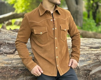 May include: A brown suede shirt with a button-down front, two chest pockets, and a classic collar. The shirt has long sleeves and gold-colored buttons. The shirt is worn over a black shirt.
