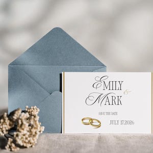 Op de afbeelding: Een witte rechthoekige save the date kaart met de namen Emily & Mark in zwarte letters, gouden zijranden en twee gouden trouwringen. De tekst luidt July 17, 2026. Een stoffig blauwe envelop staat achter de kaart.