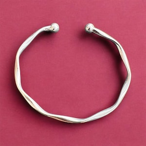 Könnte beinhalten: Ein silbernes Armreif-Armband mit einem welligen, strukturierten Design. Das Armband ist offen und hat abgerundete Enden.