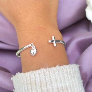 Può includere: Un braccialetto d'argento con un ciondolo a forma di cuore e un ciondolo a forma di croce. Il braccialetto è al polso.