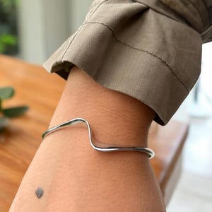 May include: A silver wave-shaped bangle bracelet on a person's wrist.