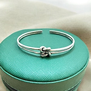 Puede incluir: Una pulsera de plata con diseño de nudo. La pulsera está hecha de dos bandas paralelas que se unen en el nudo central. La pulsera se muestra sobre una superficie verde.