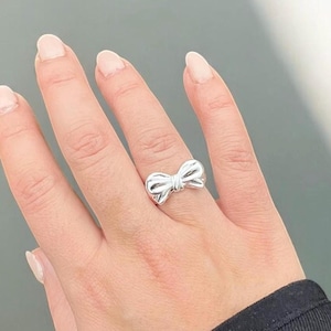 Può includere: Un anello d'argento con un design a fiocco. L'anello è indossato su un dito.