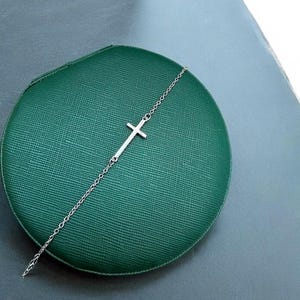 以下が含まれることがあります： 繊細なチェーンと十字架のペンダントが付いたシルバーカラーのブレスレット。ブレスレットは、質感のある濃い緑色の表面に表示されています。十字架は水平に配置されています。銀と緑のコントラストが印象的です。