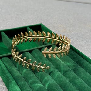 May include: A gold-colored leaf-shaped bracelet. The open cuff bracelet is made of metal and features a repeating pattern of small leaves. The bracelet is displayed on a green velvet jewelry box.