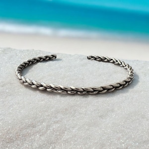 May include: A silver braided bangle bracelet. The bracelet is made of a thick, twisted wire and has a simple, elegant design.