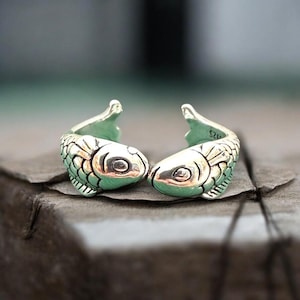 Puede incluir: Dos anillos plateados con forma de pez. Los anillos tienen escamas y aletas detalladas, con las cabezas de los peces enfrentadas. Los anillos se exhiben sobre una superficie oscura y texturizada.