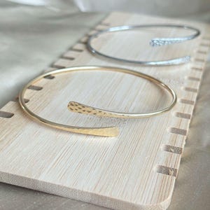 May include: Three open-ended bracelets displayed on a light-colored wooden surface. The top bracelet is silver, the middle one is gold, and the bottom one is also gold. The gold bracelets have a hammered texture on the ends.