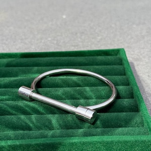 May include: A silver hinged bracelet with a cylindrical bar closure. The bracelet is displayed on a green velvet surface, against a neutral grey background. The jewellery piece is simple and modern.