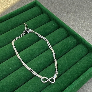 Puede incluir: Pulsera plateada de doble cadena con un símbolo de infinito y un pequeño dije de corazón. La pulsera se muestra sobre una superficie de terciopelo verde oscuro.