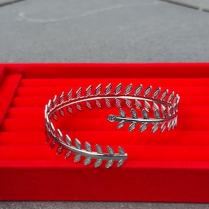 May include: Silver-toned cuff bracelet featuring a delicate leaf design. The open-ended bracelet is displayed on a red velvet surface, showcasing its intricate details and elegant style. A perfect accessory for adding a touch of nature-inspired charm.