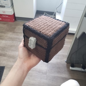Pode incluir: Um baú de crochê marrom com acabamento preto e uma alça de crochê cinza. O baú é uma réplica de um baú do Minecraft.