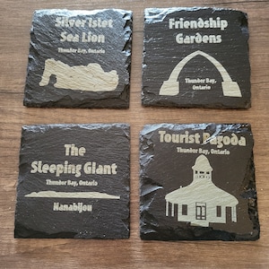 Pode incluir: Quatro  tábuas de ardósia pretas com texto e imagens brancas. As tábuas representam marcos de Thunder Bay, Ontário, Canadá. As tábuas são: Silver Islet Sea Lion, Friendship Gardens, The Sleeping Giant Nanabijou e Tourist Pagoda.