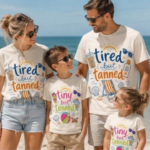 Op de afbeelding: Een gezin van vier draagt bijpassende crèmekleurige T-shirts met strandthema-afbeeldingen en tekst. De shirts zeggen "tired but tanned" en "tiny but tanned" met illustraties van zonnebrillen, een strandstoel en een drankje. Het gezin is op het strand.