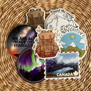 Puede incluir: Un conjunto de pegatinas con temática de viajes, con una mochila, montañas y un paisaje canadiense. Una pegatina dice "We are all made of stardust." Otra dice "Travel the world adventure."