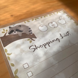 Könnte beinhalten: Einkaufszettel-Notizblock mit Aquarelldesign. Oben links ist ein schlafender Waschbär auf einem Ast abgebildet. Die Worte "Shopping List" sind kursiv geschrieben. Enthält Apfelillustrationen und Kontrollkästchen.