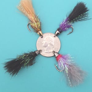 May include: Four colorful fishing flies arranged around a quarter. The flies have black, purple, gray, and tan fur tails with silver tinsel accents. Each fly has a silver hook and a black metal head.