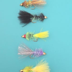 May include: Five fishing flies arranged vertically. The top fly is tan with iridescent fibers. The second is black with silver tinsel. The third is yellow and green with a red head. The fourth is purple with silver fibers. The bottom fly is yellow and green.